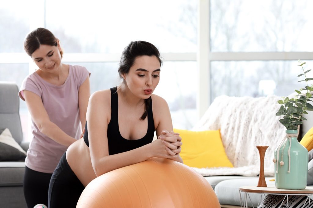 A pregnant woman in a black sports bra and leggings sitting on a large orange birthing ball while a doula or childbirth educator gently rubs her back in a brightly lit living room.