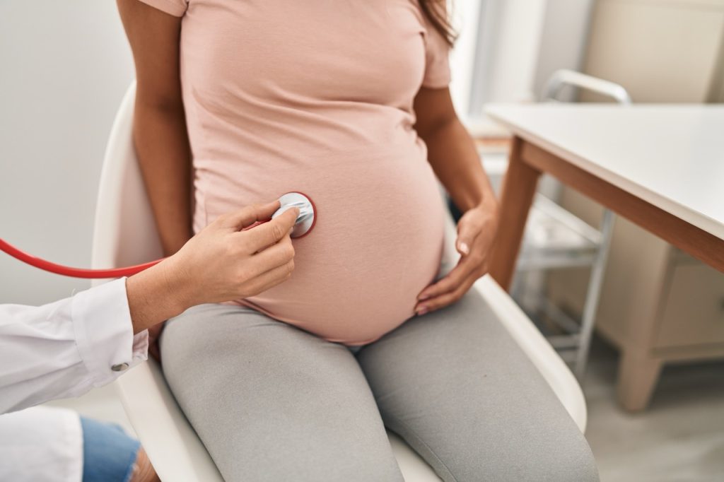 A close-up view of a healthcare provider's hands using a stethoscope to examine the belly of a pregnant woman during a clinical checkup.