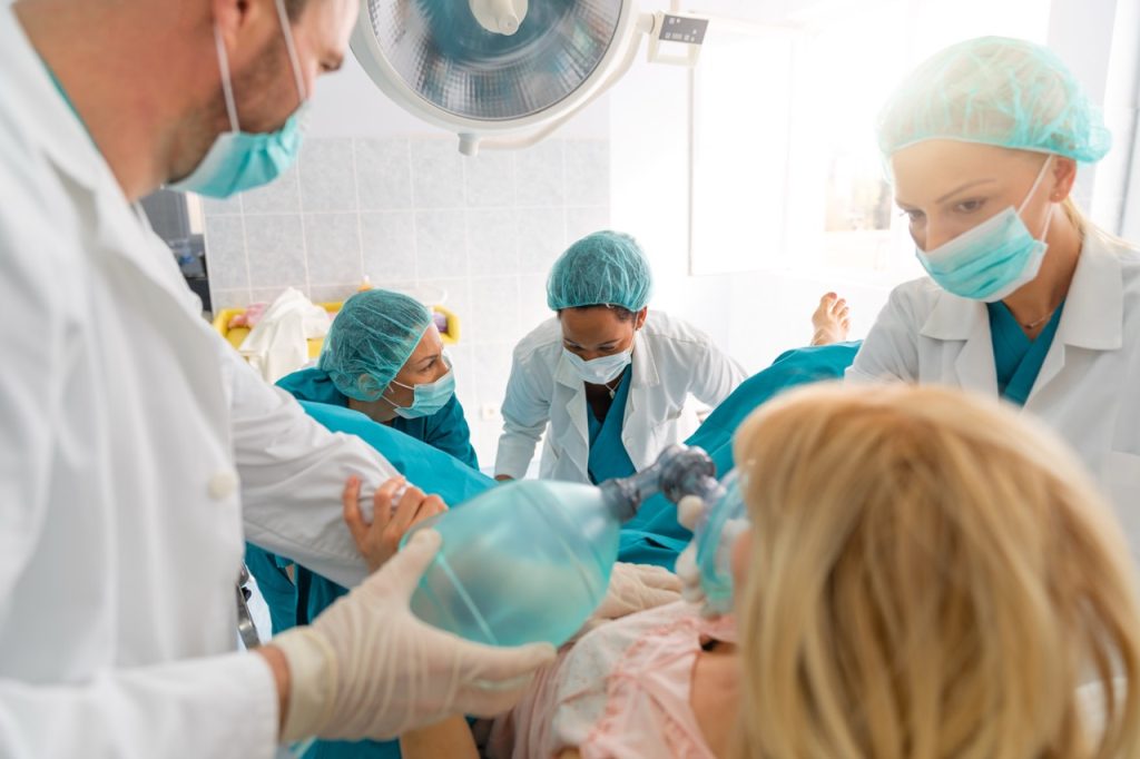 A medical team of doctors and nurses in scrubs and face masks working together around a hospital bed to support and encourage a pregnant woman during childbirth in a delivery room.