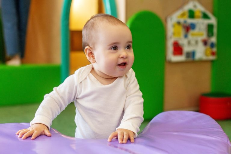 Smiling baby in a white onesie standing while holding onto colorful soft play equipment.